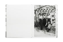 Load image into Gallery viewer, Spread from book, no image on the left. On the right, b/w photo of four Chicana women, three standing and one crouching and holding an infant. All four looking at the camera. The three standing have highlighted hair, two have their hands clasped together. All three standing have dark pants or jeans and white tops of various styles with bare arms. Behind them, a fence painted with large WF graffiti and other messages.