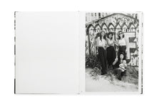 Load image into Gallery viewer, Spread from book, no image on the left. On the right, b/w photo of four Chicana women, three standing and one crouching and holding an infant. All four looking at the camera. The three standing have highlighted hair, two have their hands clasped together. All three standing have dark pants or jeans and white tops of various styles with bare arms. Behind them, a fence painted with large WF graffiti and other messages. 

