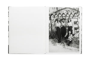 Spread from book, no image on the left. On the right, b/w photo of four Chicana women, three standing and one crouching and holding an infant. All four looking at the camera. The three standing have highlighted hair, two have their hands clasped together. All three standing have dark pants or jeans and white tops of various styles with bare arms. Behind them, a fence painted with large WF graffiti and other messages. 