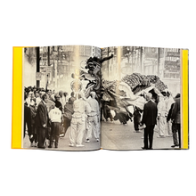 Load image into Gallery viewer, Open book showing a black and white photograph of a parade in Vancouver&#39;s Chinatown with onlookers watching lion dancers perform.
